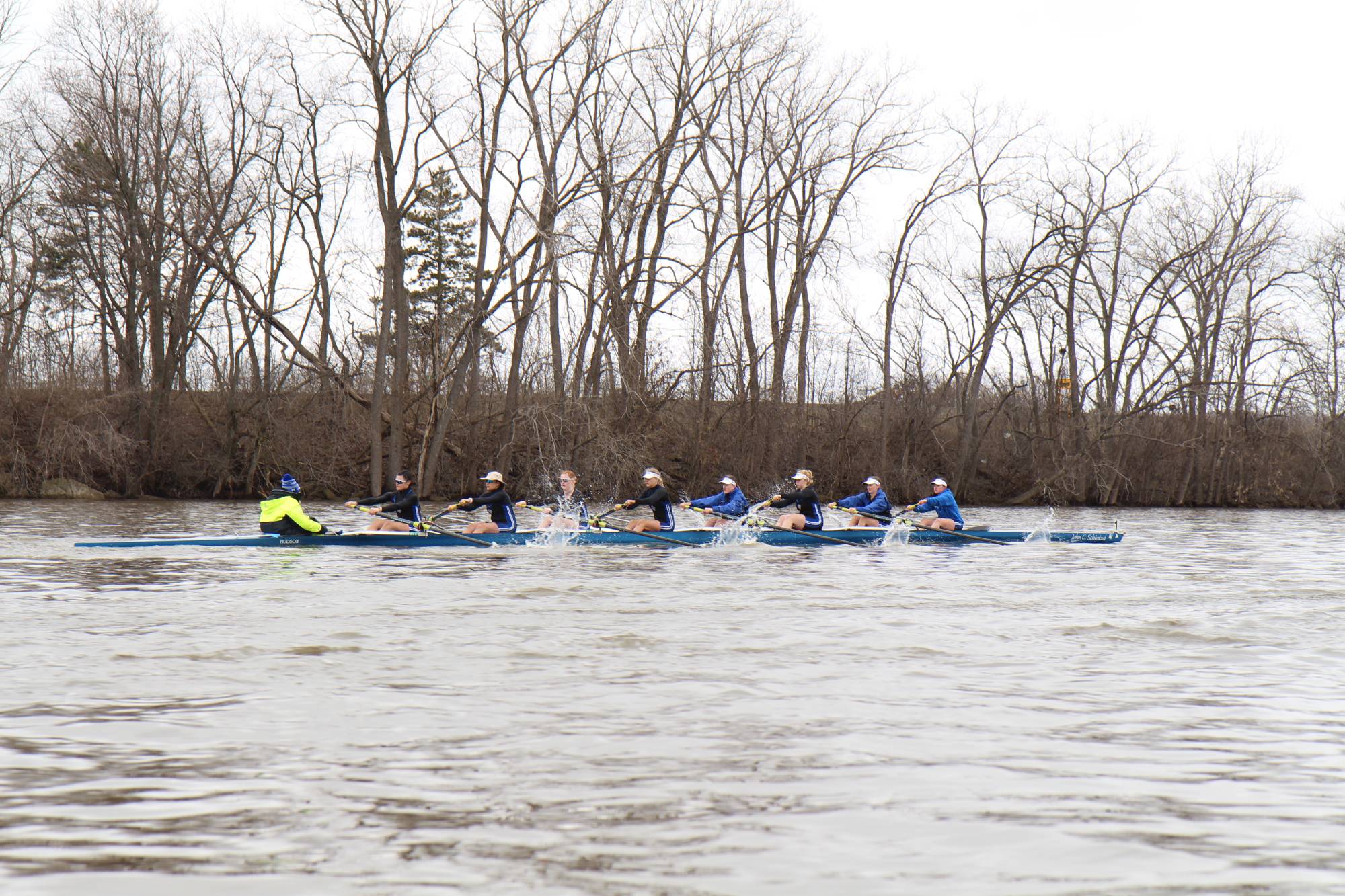 8 womens rowing during an overcast day on a river with trees with no leaves in the background